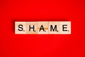 Photo of Scrabble game tiles spelling out the word 'shame' on a bright red background.